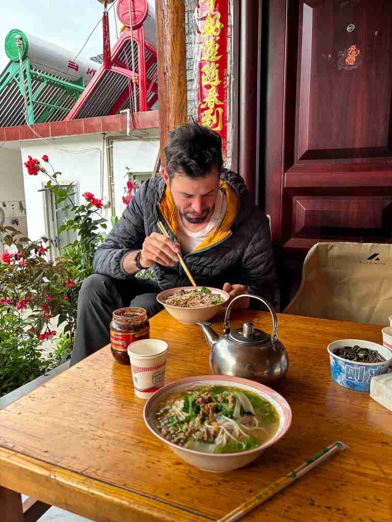 Traveler enjoying delicious home-cooked pork noodle soup at wooden table in front of traditional red Chinese building with flowers and mountain view