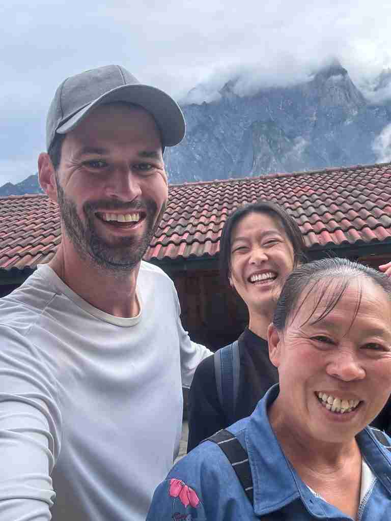 Corry and Ling smiling with their local host at the family homestay in Halfway, Tiger Leaping Gorge, with traditional red-tiled roof building and misty mountains in background