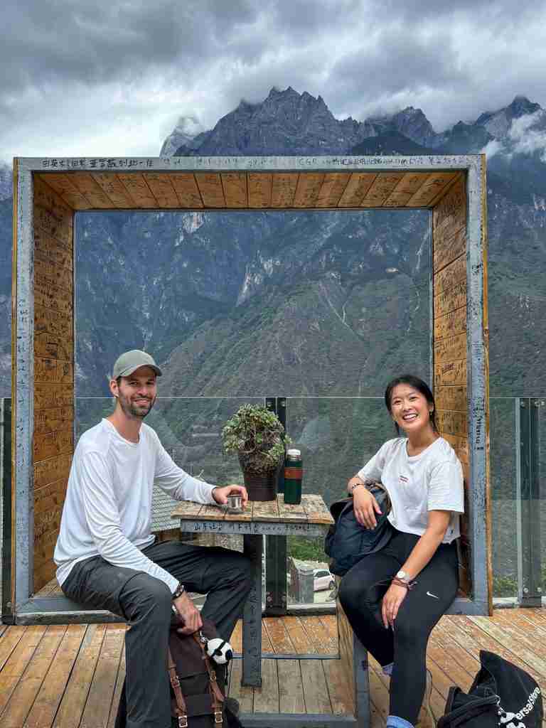 Corry and Ling sitting at scenic wooden viewpoint frame overlooking Tiger Leaping Gorge with dramatic misty mountains in background