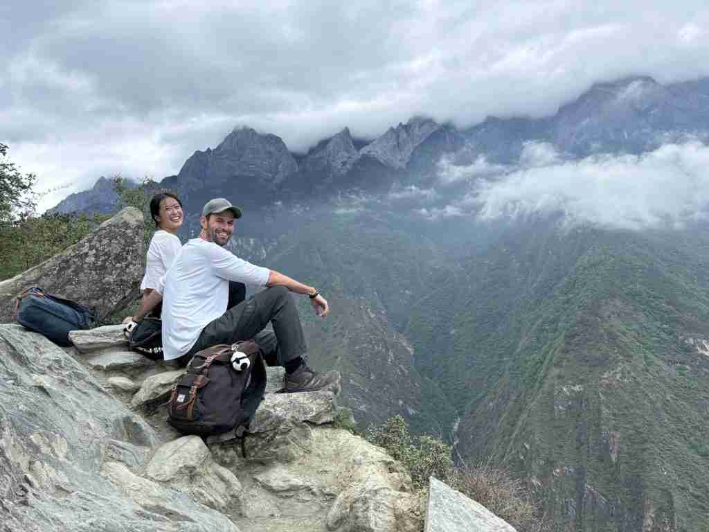 Corry and Ling sitting on large rocky outcrop overlooking Tiger Leaping Gorge with dramatic misty mountains, deep gorge, and green valleys in background