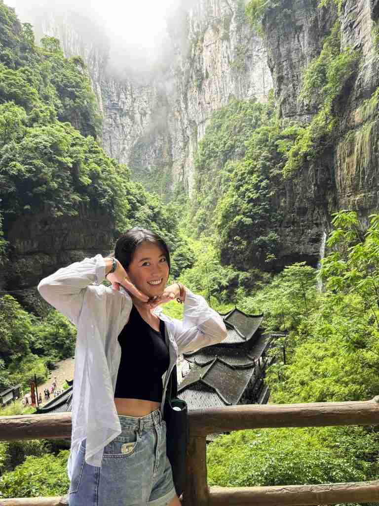 A smiling traveler posing at the Wulong Karst National Park in Chongqing with the traditional Tianlong Outpost and limestone cliffs in the background.