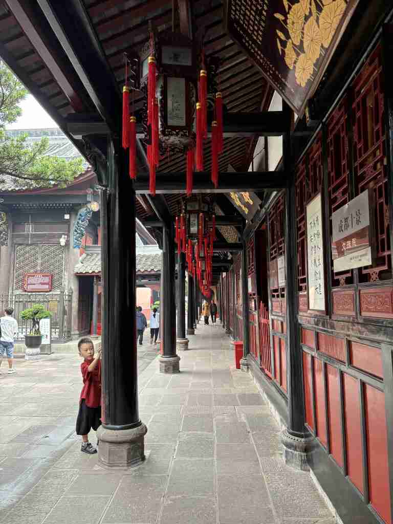 A view of the traditional covered walkway at Wenshu Monastery, a serene Buddhist temple in Chengdu. A long, covered walkway with ornate red walls, dark wooden pillars, and traditional red lanterns hanging from the ceiling at Wenshu Monastery in Chengdu. A young child in a red shirt is visible peeking from behind one of the pillars on the left, adding a sense of life to the historical scene.