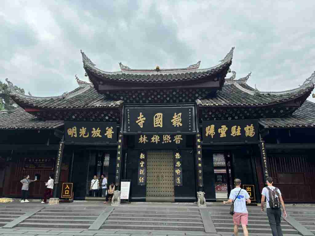 The main gate of Baoguo Temple in Chengdu, a famous Buddhist temple with traditional Chinese architecture. A grand, wide-shot photograph of the traditional Chinese architectural entrance of Baoguo Temple in Chengdu under a cloudy sky, featuring ornate roofs, large black doors with golden characters, and stone steps leading up to the entrance where several visitors are entering.