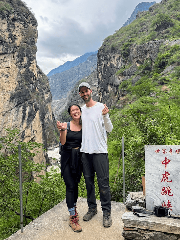 A happy couple hiking in China stands at a viewpoint overlooking the Jinsha River. The image highlights how to get from Lijiang to Tiger Leaping Gorge for scenic trekking.