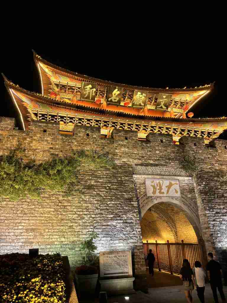 A night photograph of the South Gate of Dali Ancient City, beautifully illuminated with warm golden lights, highlighting the traditional Chinese architecture and ornate roof. The archway is glowing, with a few people walking in the foreground, capturing the gate's peaceful charm after dark.