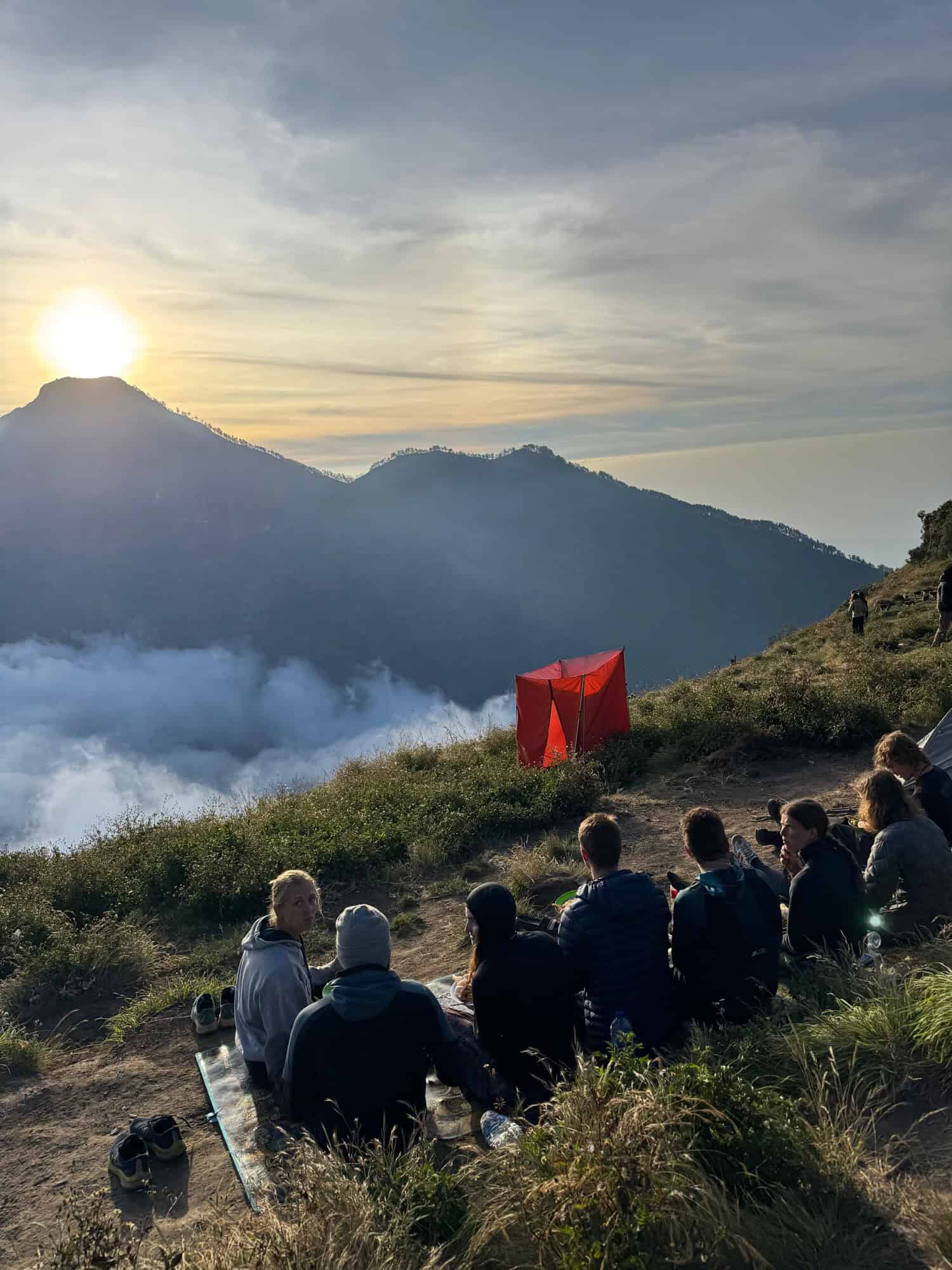 Hikers sit on a cliffside at sunset, watching the sun descend over Mt Rinjani's landscape, with a vibrant sky and Silhouetted tents in the distance.