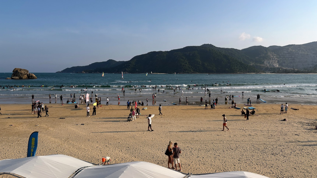Beginner surfers practicing in the shallow, gentle waves of Houhai Bay with mountains in the background.