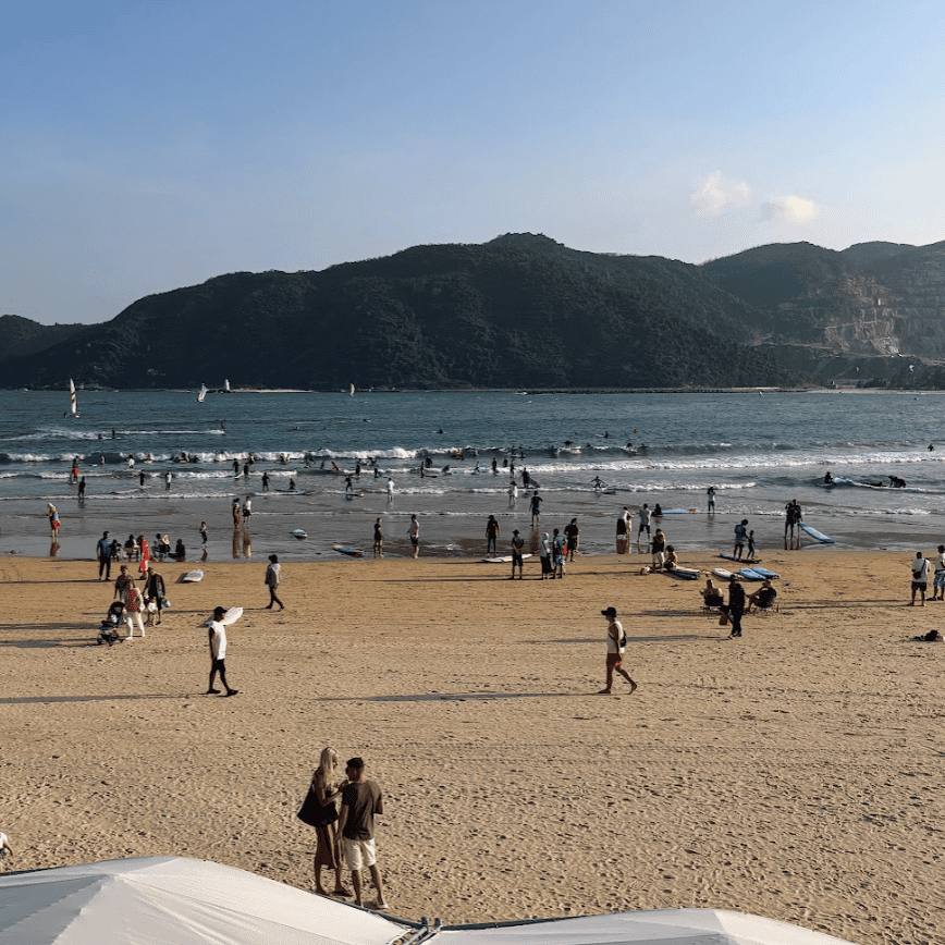 Beginner surfers practicing in the shallow, gentle waves of Houhai Bay with mountains in the background.