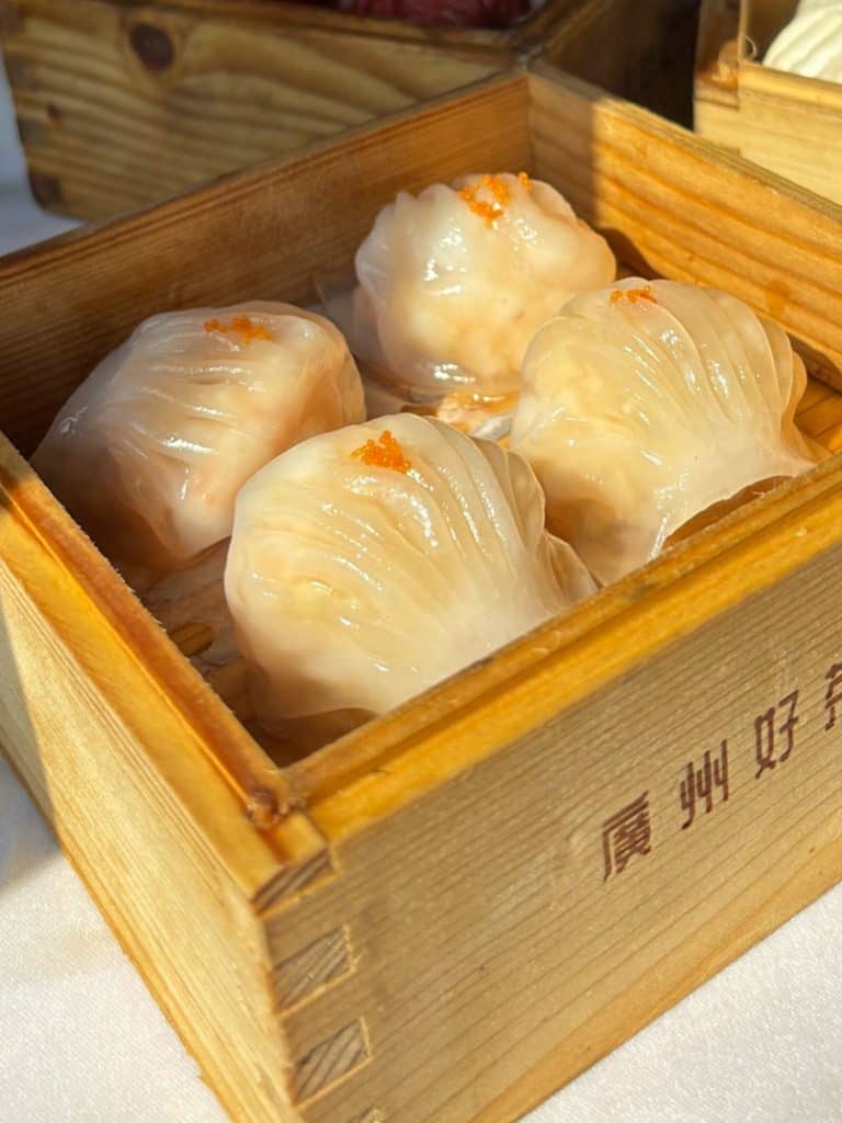 Four translucent shrimp dumplings topped with orange roe in a wooden steamer basket.
