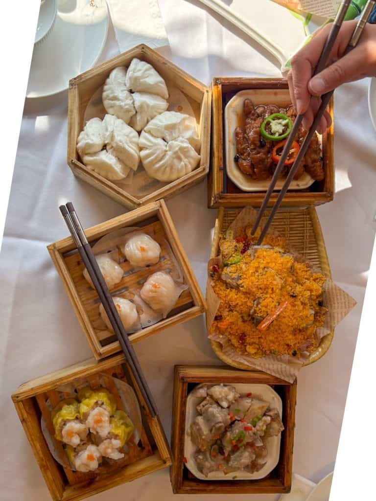 An array of wooden steamers containing chicken feet, pork ribs, buns, and dumplings on a white table.