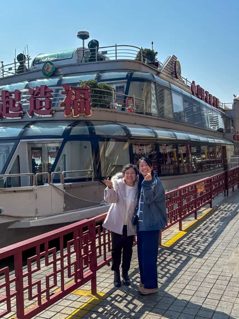 Two women smiling in front of a large riverboat with Chinese characters on the side.