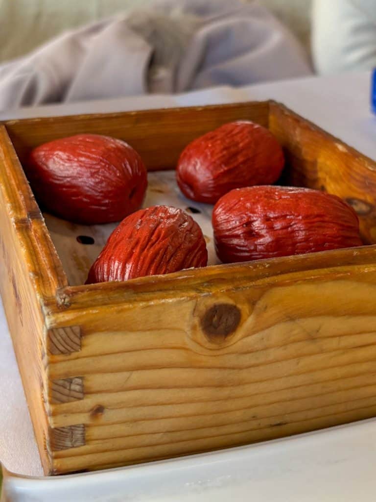 Four dark red, wrinkled jujube cakes served in a square wooden steamer.