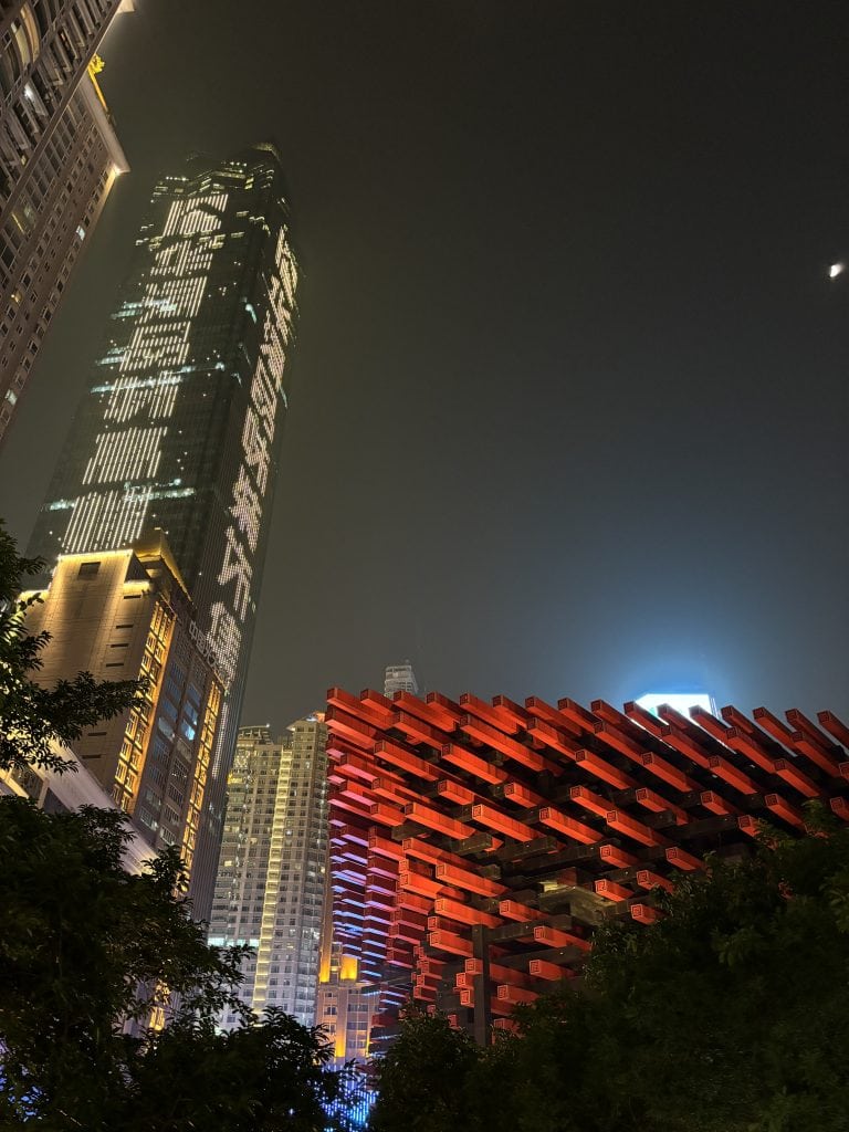 The red interlocking beam architecture of the Chongqing Guotai Arts Center at night with a glowing skyscraper in the background.