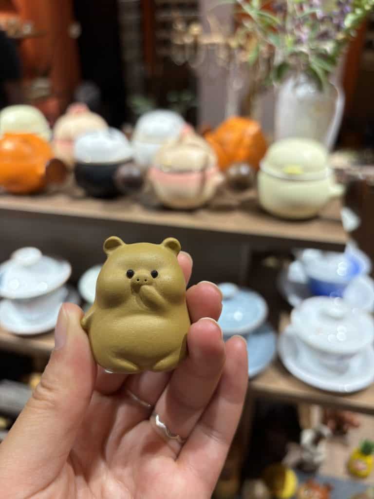 A hand holding a small, brown ceramic bear-shaped tea pet in a Chongqing boutique shop with blurred tea sets in the background.