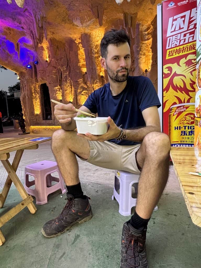 A traveler sitting on a plastic stool and eating street food with chopsticks outside an illuminated cave entrance in Chongqing.