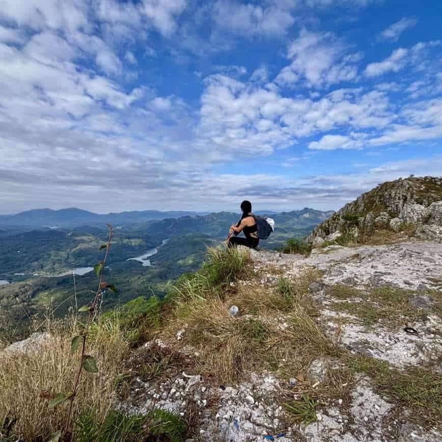A female hiker sitting on a rocky peak at Baishi Ridge in Sanya, Hainan, overlooking a winding river and lush green valley under a blue sky.