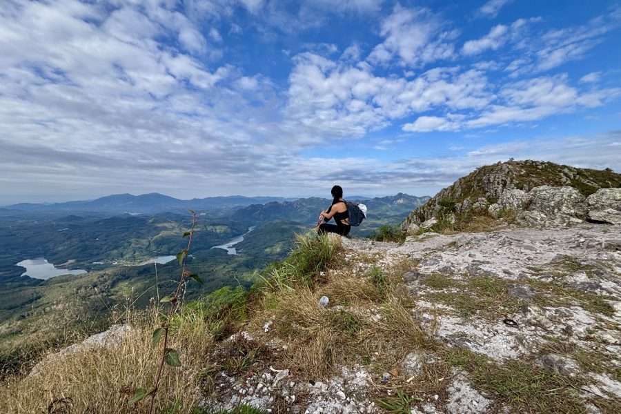 A female hiker sitting on a rocky peak at Baishi Ridge in Sanya, Hainan, overlooking a winding river and lush green valley under a blue sky.