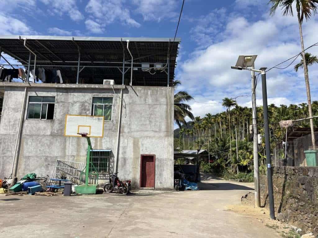 A concrete building with a basketball hoop in a rural Hainan village surrounded by palm trees and mountains.