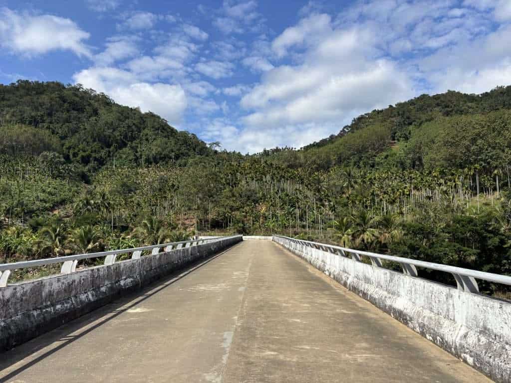 A long concrete bridge leading toward lush tropical mountains and palm oil plantations under a cloudy blue sky in Hainan.
