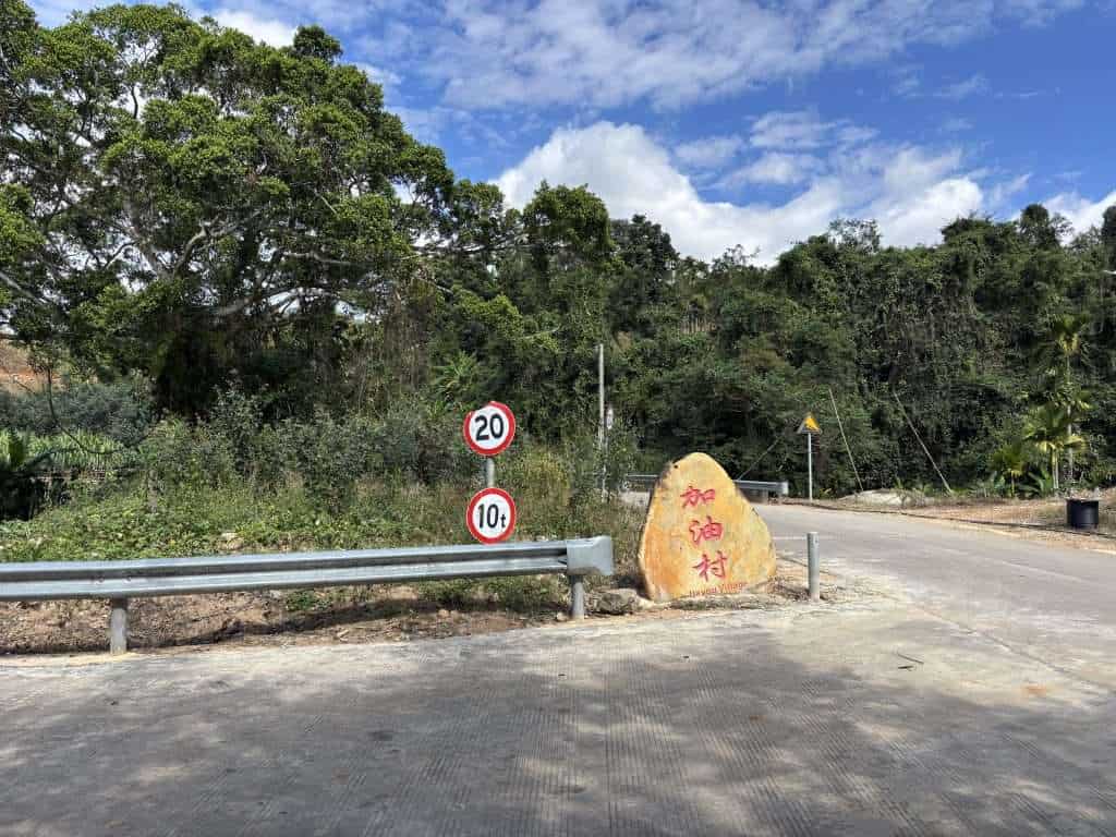 Entrance to Jiayou Village near Baishi Ridge in Sanya with a large stone marker featuring red Chinese characters and road speed limit signs.