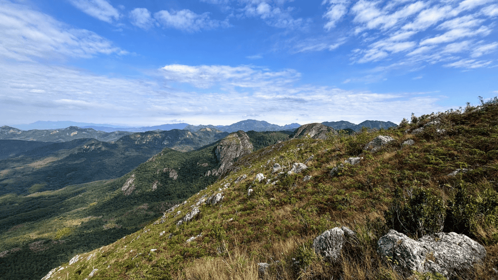 Panoramic landscape of the rocky Baishi Ridge trail showing green mountain peaks and tropical forests under a bright blue sky in Sanya, Hainan.