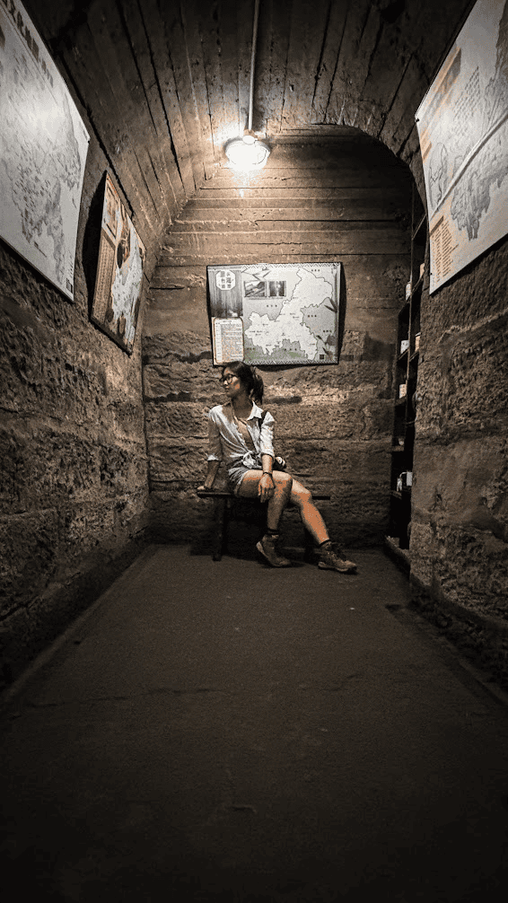 A woman sitting on a wooden bench inside a stone-walled underground air raid shelter tunnel repurposed as a bookstore in Chongqing, featuring historical maps and warm overhead lighting.