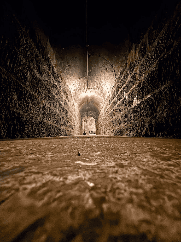 A low-angle, long-distance view of a dimly lit, arched stone tunnel inside a repurposed air raid shelter in Chongqing, China, showing the textured rock walls and industrial lighting.