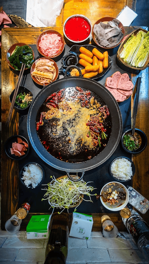 Top-down view of a spicy Sichuan-style hotpot table spread with a bubbling dark broth, surrounded by side dishes like tripe, bean sprouts, and sliced meats.