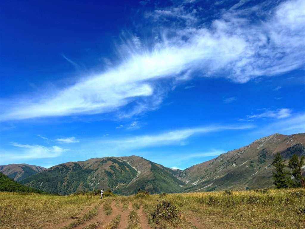 Wide valley view from the horse riding route to Beauty Peak in Hemu Village, green mountains under a bright blue sky with wispy clouds