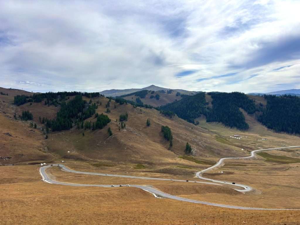 Winding sealed road through golden hills on the Ahe Highway, with scattered yurts visible in the distance