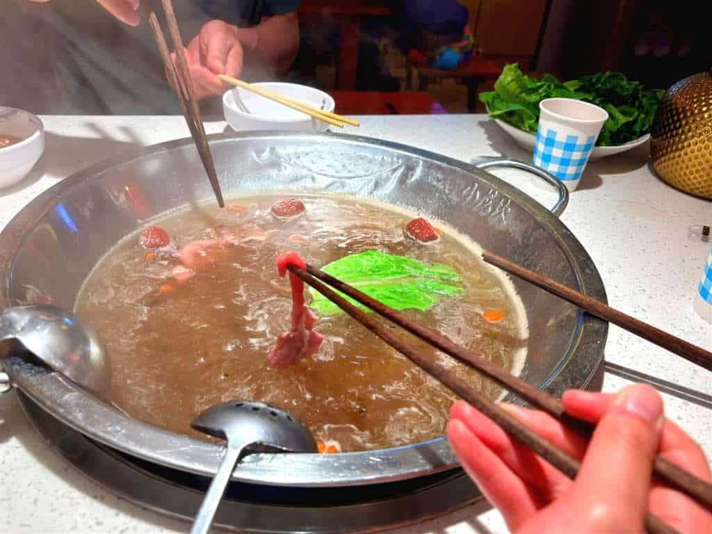Hotpot cooking in a shared pot at 小龙坎火锅 in Hemu Village, chopsticks lowering meat and vegetables into the broth